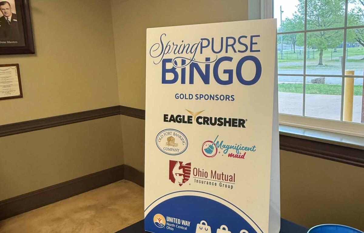 Tall sign reading 'SpringPurse BINGO' with sponsor logos on a table inside a room near a window.