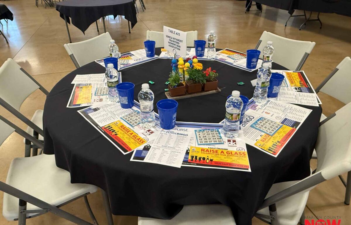 Table #25 set for a banquet with a black cloth, water bottles, blue cups, flyers, and a flower centerpiece in a large event hall.