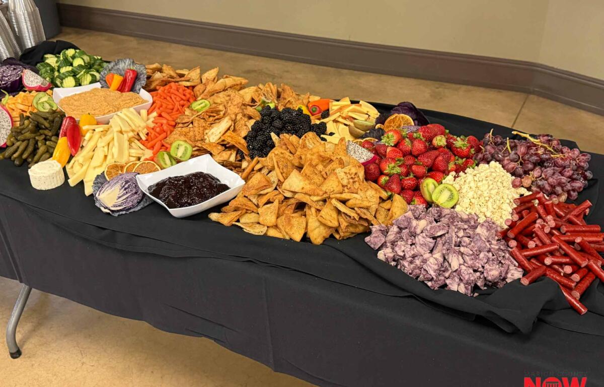 Long buffet table covered with a black cloth displaying a colorful charcuterie spread: crackers, tortilla chips, cheeses, sliced veggies, fruits like strawberries and kiwis, and bowls of dips, with a red NOW logo in the corner.