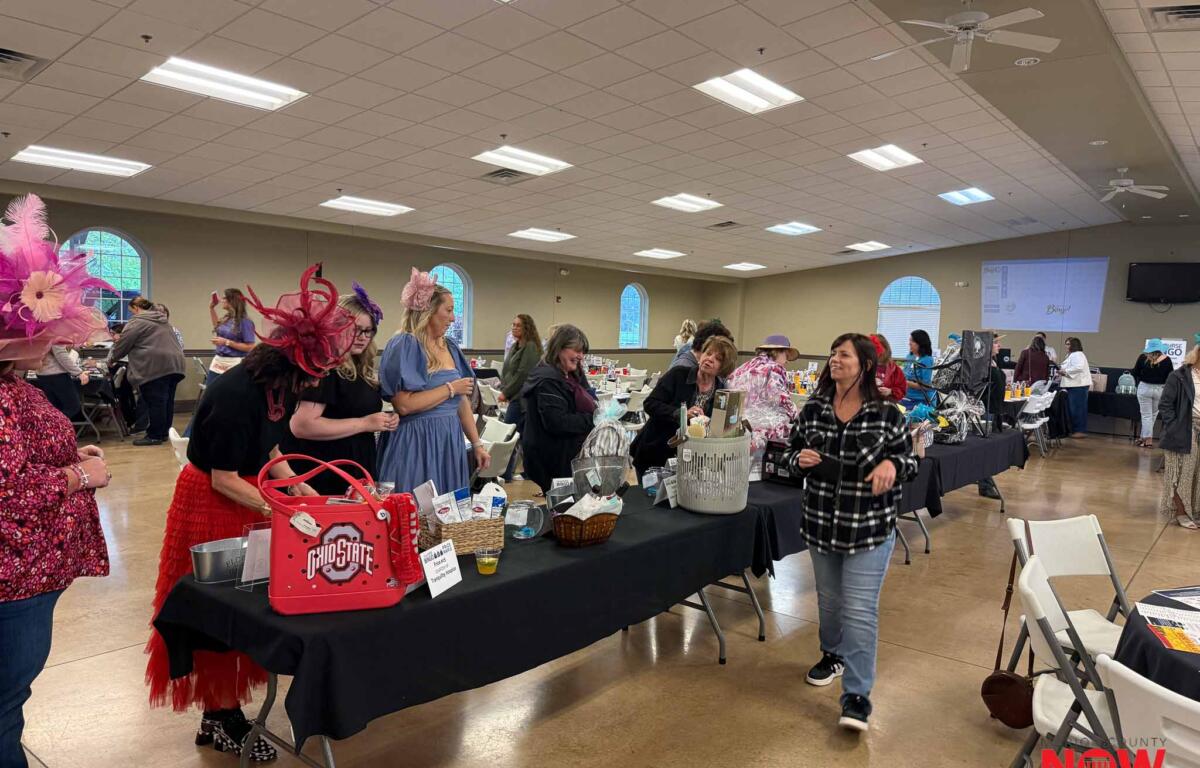 Busy indoor event in a hall with long tables displaying gift baskets and prizes; people browse and chat in colorful outfits and hats.