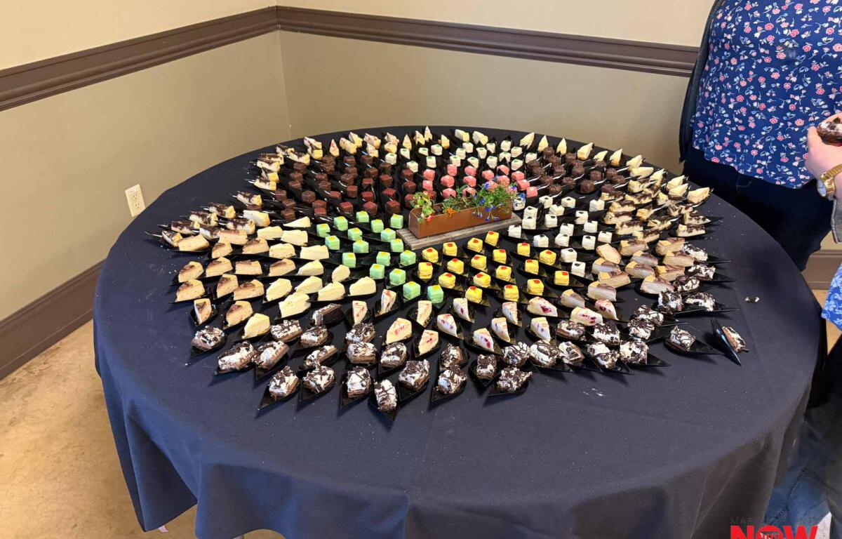 Table covered with a dark cloth displays a circular arrangement of bite-sized desserts on skewers, with a rectangular planter centerpiece in the middle and a person in a blue floral shirt partially visible on the right.