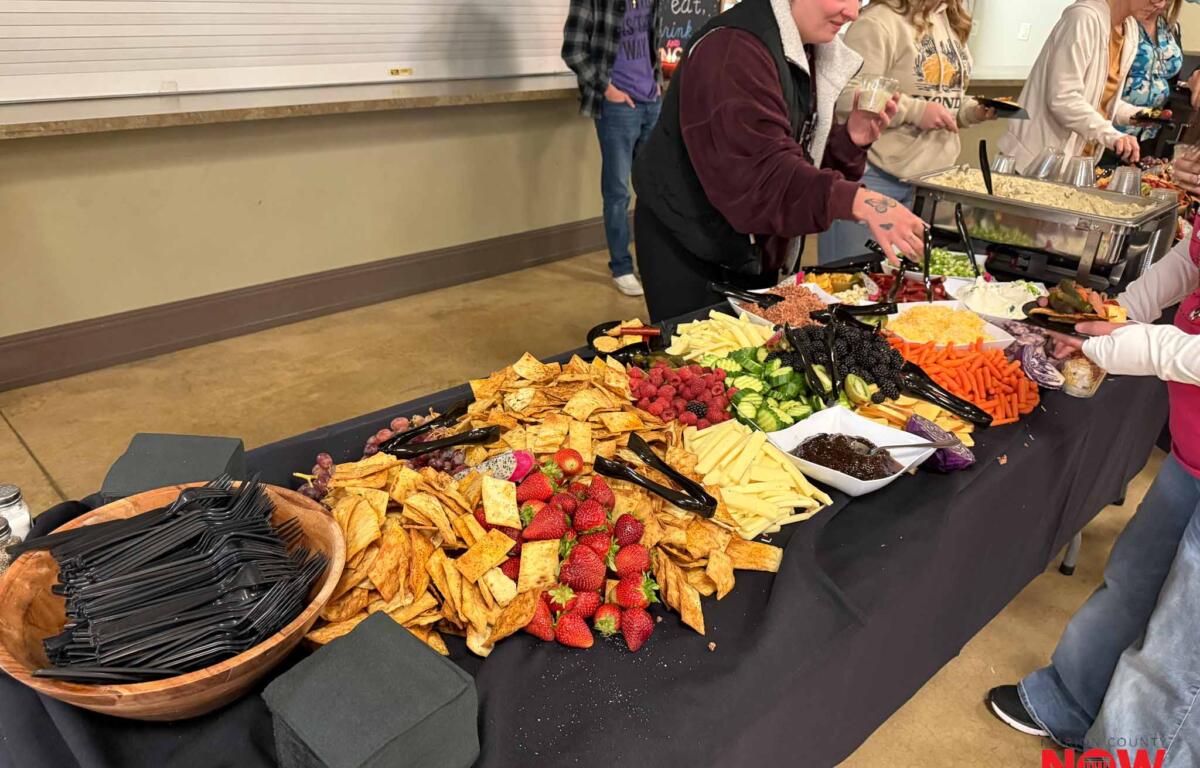 Buffet table with tortilla chips, fruit, cheese, and dips as several people serve themselves at a casual gathering.