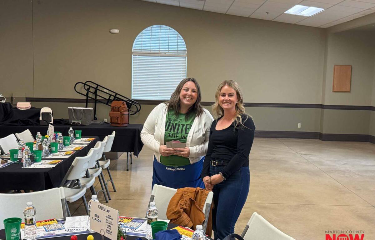 Two smiling women posing for a photo at a United Way event in a banquet hall, with tables set with water bottles, name tents, and colorful game pieces in the foreground.