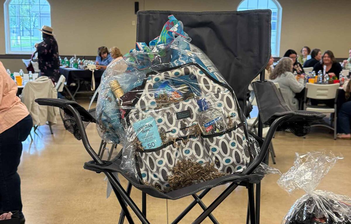 Prize raffle display in a banquet hall: a large wrapped gift basket on a folding chair with raffle signs and attendees at tables in the background