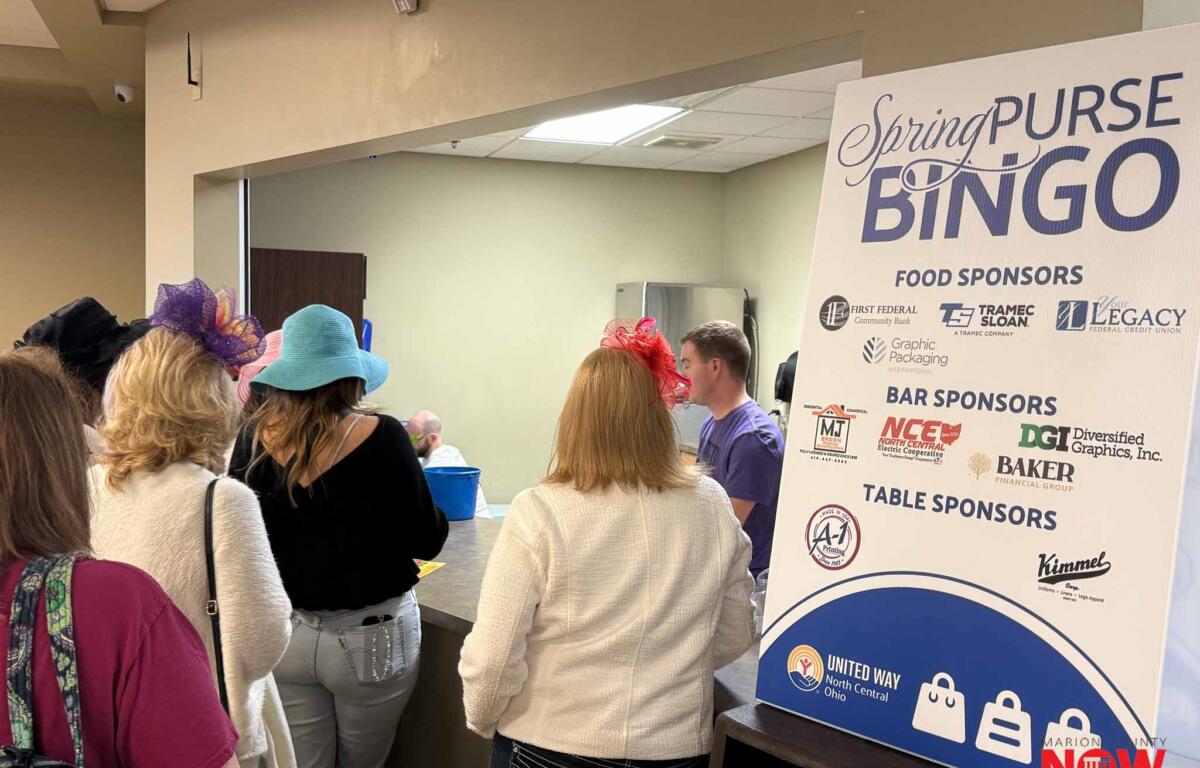 People gathered at an indoor bingo event beside a large sponsor board labeled Spring Purse Bingo with logos beneath, viewed from the back.