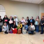 Group of women posing with assorted handbags indoors, some kneeling in front; Marion County NOW logo bottom right.