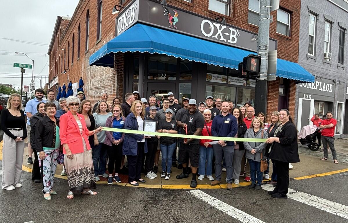 Group of people gathered outside Ox-B's restaurant for a ribbon‑cutting ceremony, holding a yellow ribbon across the storefront.