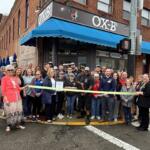 Group of people gathered outside Ox-B's restaurant for a ribbon‑cutting ceremony, holding a yellow ribbon across the storefront.