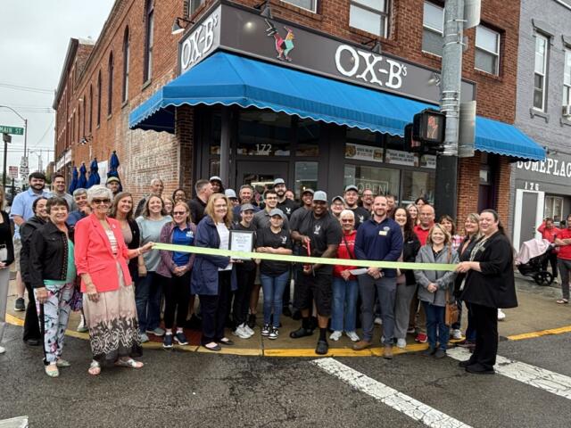 Group of people gathered outside Ox-B's restaurant for a ribbon‑cutting ceremony, holding a yellow ribbon across the storefront.