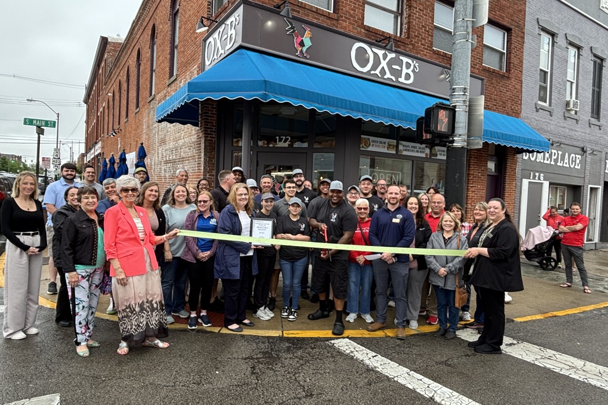 Group of people gathered outside Ox-B's restaurant for a ribbon‑cutting ceremony, holding a yellow ribbon across the storefront.