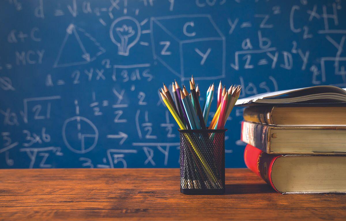 Pencil cup with colorful pencils beside a stack of books on a wooden desk, with a blue chalkboard full of math formulas in the background.