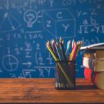 Pencil cup with colorful pencils beside a stack of books on a wooden desk, with a blue chalkboard full of math formulas in the background.