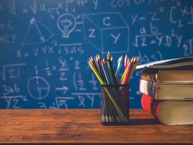 Pencil cup with colorful pencils beside a stack of books on a wooden desk, with a blue chalkboard full of math formulas in the background.