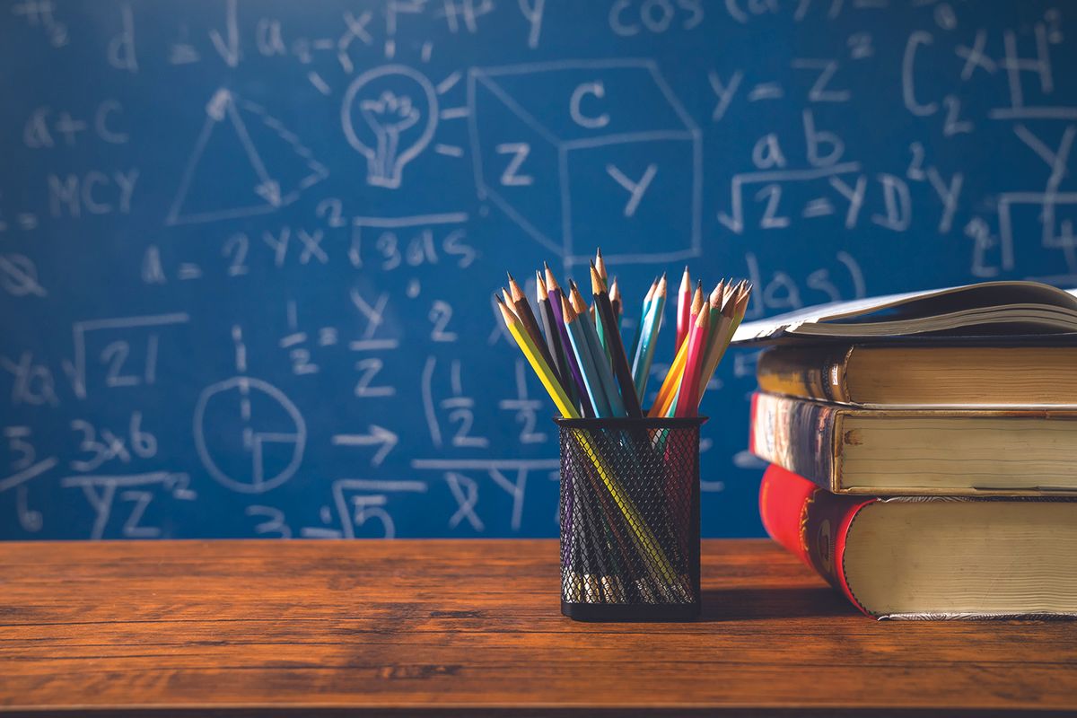 Pencil cup with colorful pencils beside a stack of books on a wooden desk, with a blue chalkboard full of math formulas in the background.