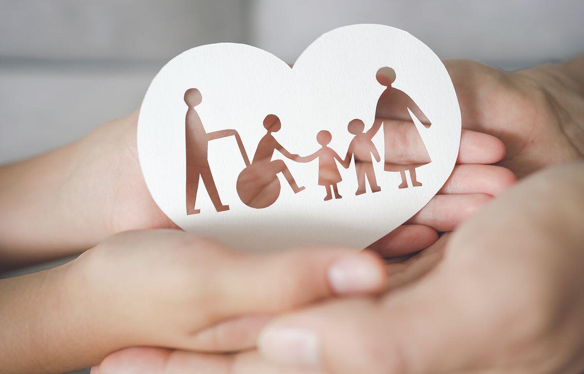 Heart-shaped card with brown cutout of a family: a person in a wheelchair, two children, and two adults holding hands, cradled in protective hands.
