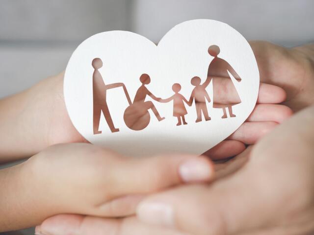 Heart-shaped card with brown cutout of a family: a person in a wheelchair, two children, and two adults holding hands, cradled in protective hands.