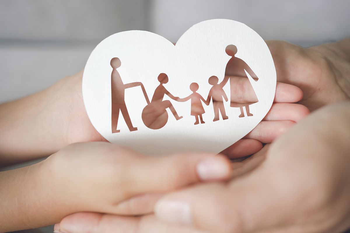 Heart-shaped card with brown cutout of a family: a person in a wheelchair, two children, and two adults holding hands, cradled in protective hands.