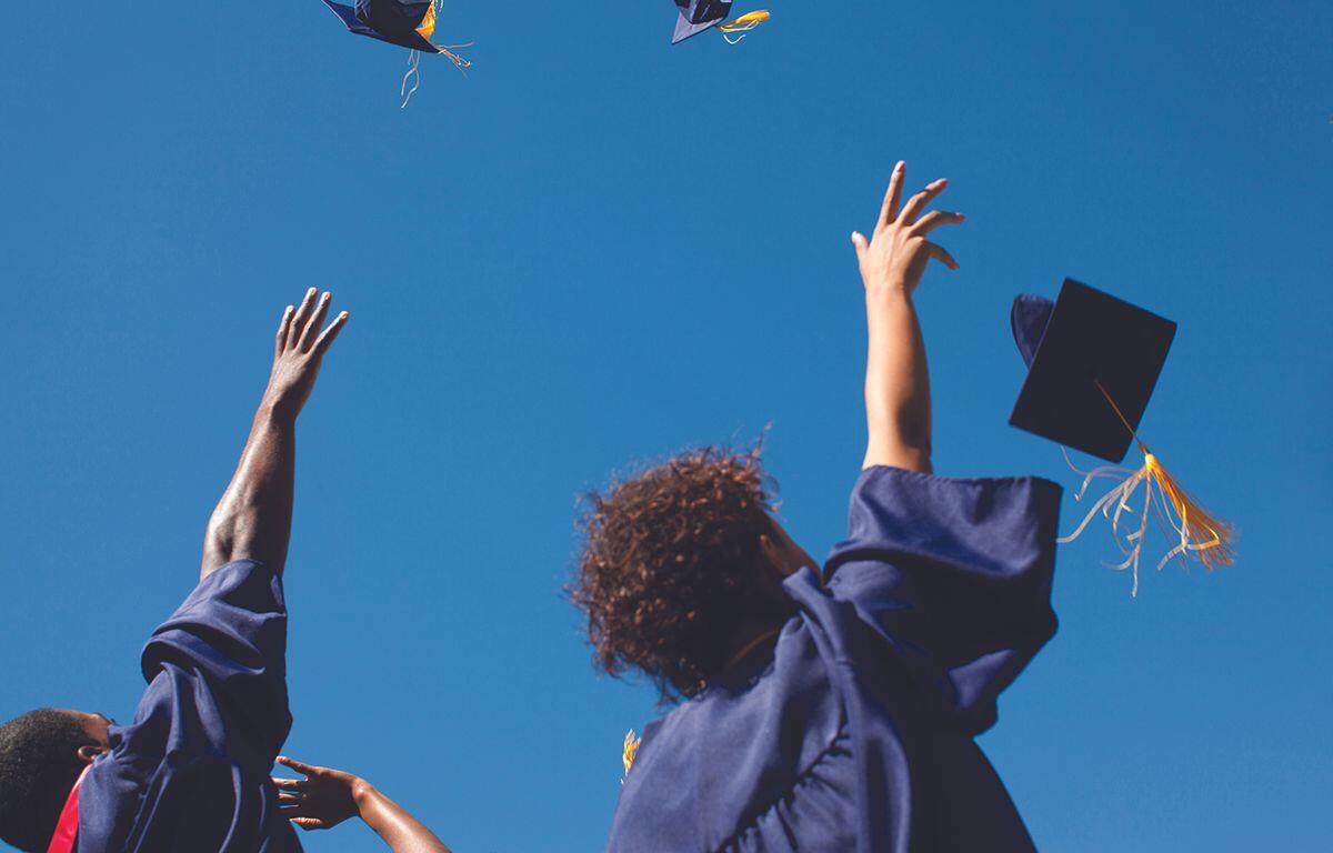 Group of graduates in blue gowns tossing their caps in the air against a clear blue sky, celebrating graduation.
