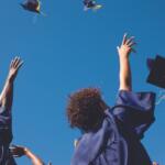 Group of graduates in blue gowns tossing their caps in the air against a clear blue sky, celebrating graduation.