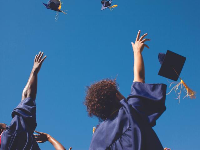 Group of graduates in blue gowns tossing their caps in the air against a clear blue sky, celebrating graduation.
