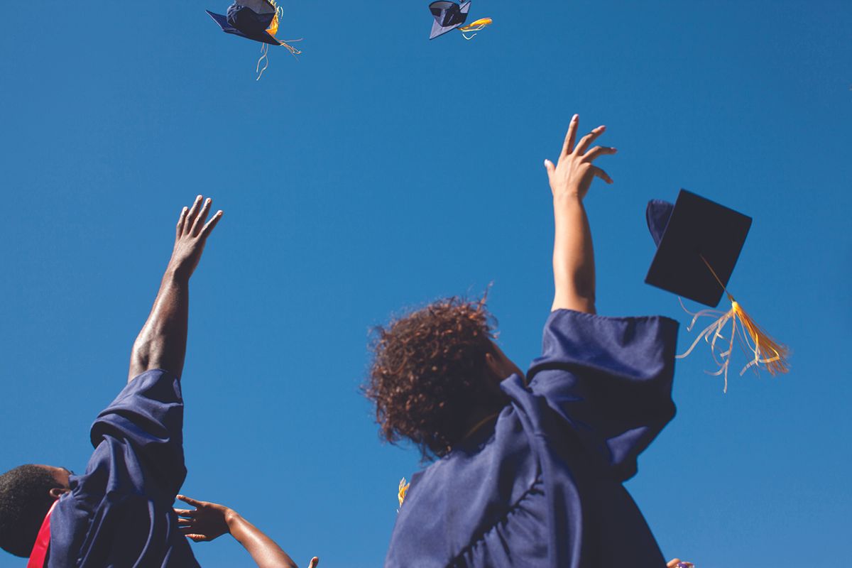 Group of graduates in blue gowns tossing their caps in the air against a clear blue sky, celebrating graduation.