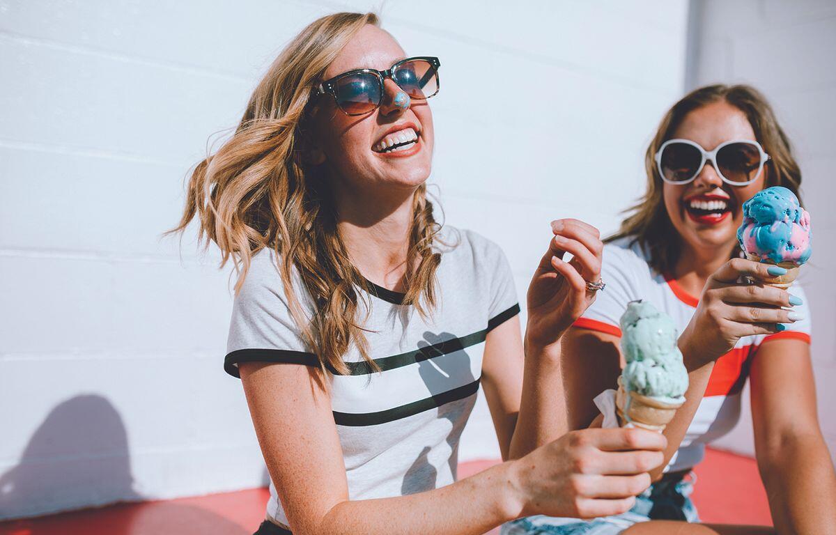 Two smiling women wearing sunglasses share colorful ice cream cones outdoors on a sunny day.