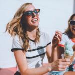 Two smiling women wearing sunglasses share colorful ice cream cones outdoors on a sunny day.