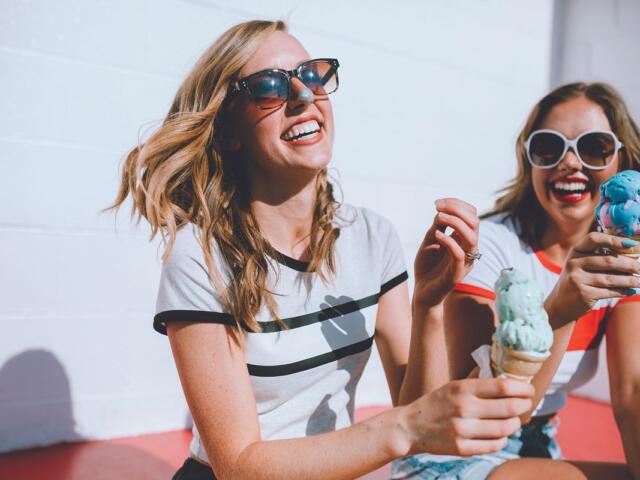 Two smiling women wearing sunglasses share colorful ice cream cones outdoors on a sunny day.