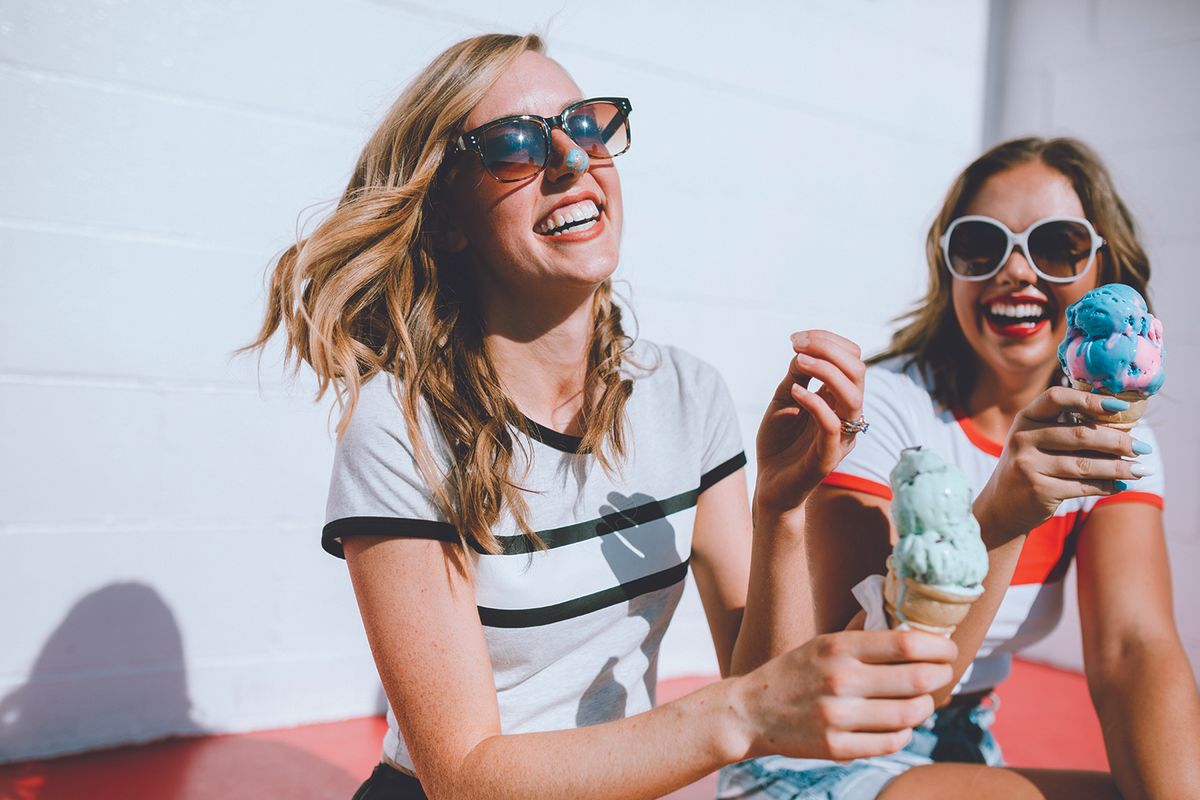 Two smiling women wearing sunglasses share colorful ice cream cones outdoors on a sunny day.