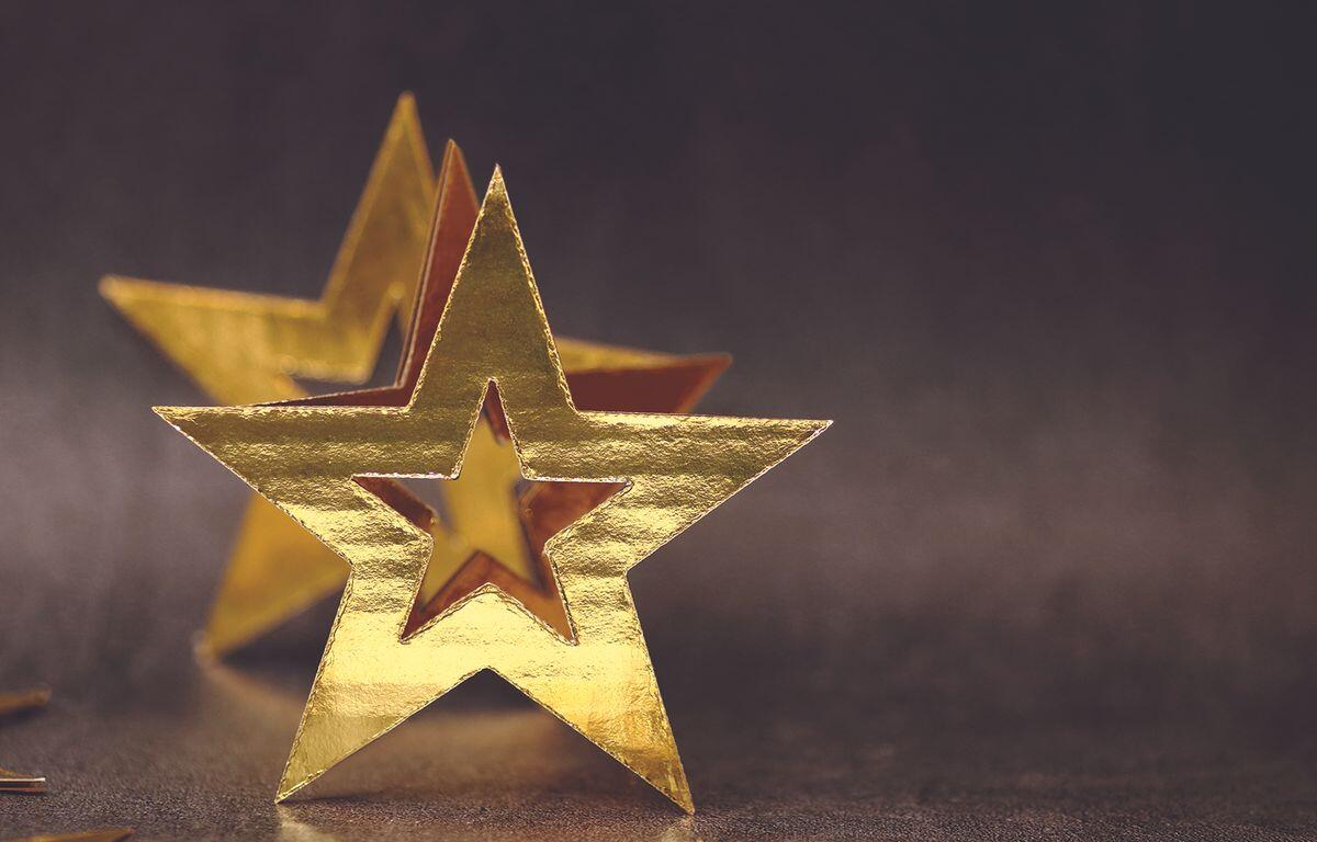 Gold foil star decorations on a dark, shallowly blurred background, with one star in front and another behind.