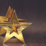 Gold foil star decorations on a dark, shallowly blurred background, with one star in front and another behind.