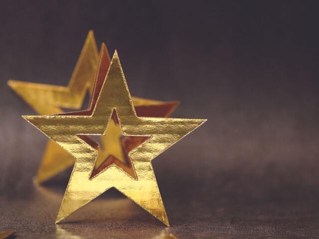 Gold foil star decorations on a dark, shallowly blurred background, with one star in front and another behind.