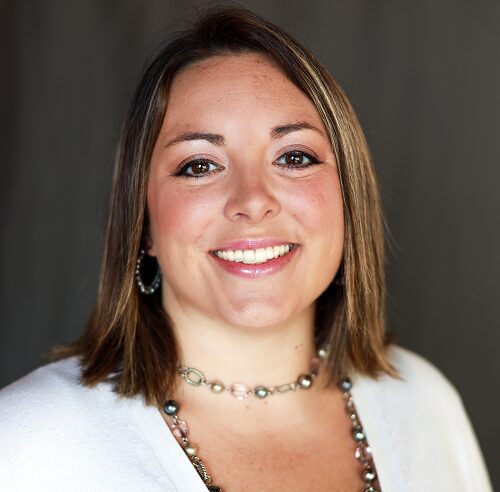 Smiling woman with shoulder-length brown hair wearing a white cardigan and beaded necklace (head-and-shoulders portrait)