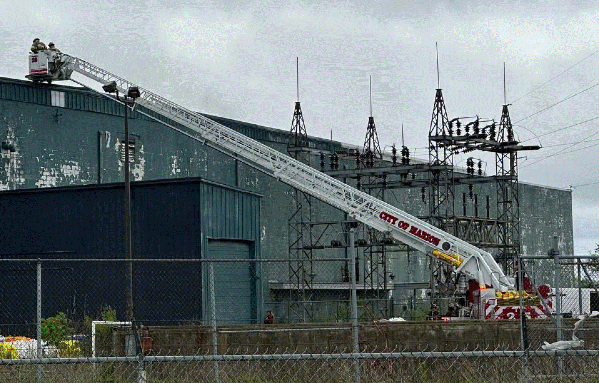 Aerial ladder truck with its zigzag boom extended toward an industrial building, firefighters on the platform at the top.