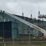 Aerial ladder truck with its zigzag boom extended toward an industrial building, firefighters on the platform at the top.
