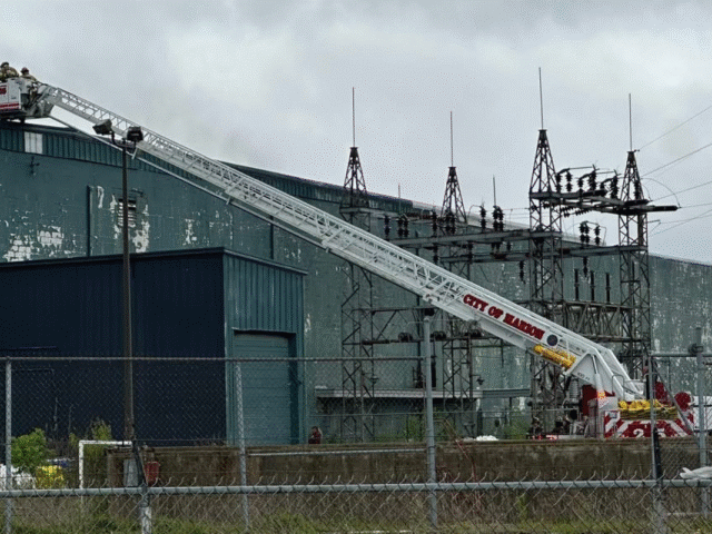 Aerial ladder truck with its zigzag boom extended toward an industrial building, firefighters on the platform at the top.