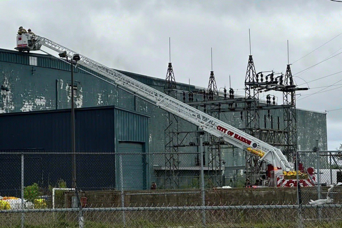 Aerial ladder truck with its zigzag boom extended toward an industrial building, firefighters on the platform at the top.