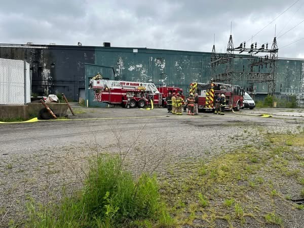 Firefighters in gear stand near two red fire trucks with hoses outside a weathered industrial building under cloudy skies.