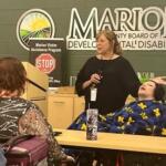 Woman with a microphone speaks to a small audience in a meeting room; a person in a wheelchair sits at the front, wrapped in a blue and yellow blanket, near a Marion banner.