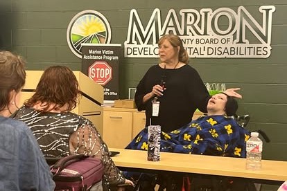 Woman with a microphone speaks to a small audience in a meeting room; a person in a wheelchair sits at the front, wrapped in a blue and yellow blanket, near a Marion banner.