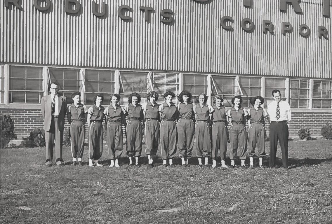 Group of factory workers in matching coveralls standing in a line outside a brick building with a 'PRODUCTS CORP' sign above. Two men in suits flank the line at each end.