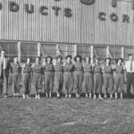Group of factory workers in matching coveralls standing in a line outside a brick building with a 'PRODUCTS CORP' sign above. Two men in suits flank the line at each end.