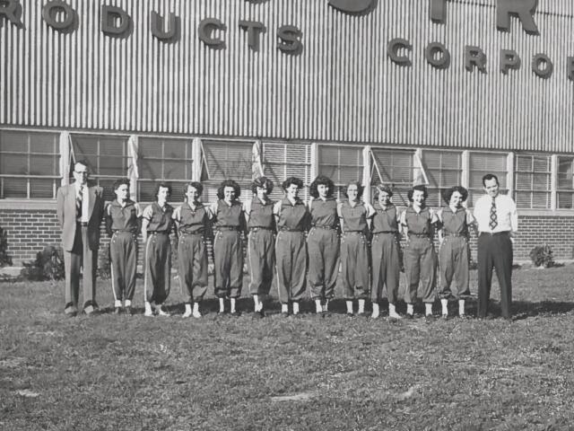 Group of factory workers in matching coveralls standing in a line outside a brick building with a 'PRODUCTS CORP' sign above. Two men in suits flank the line at each end.