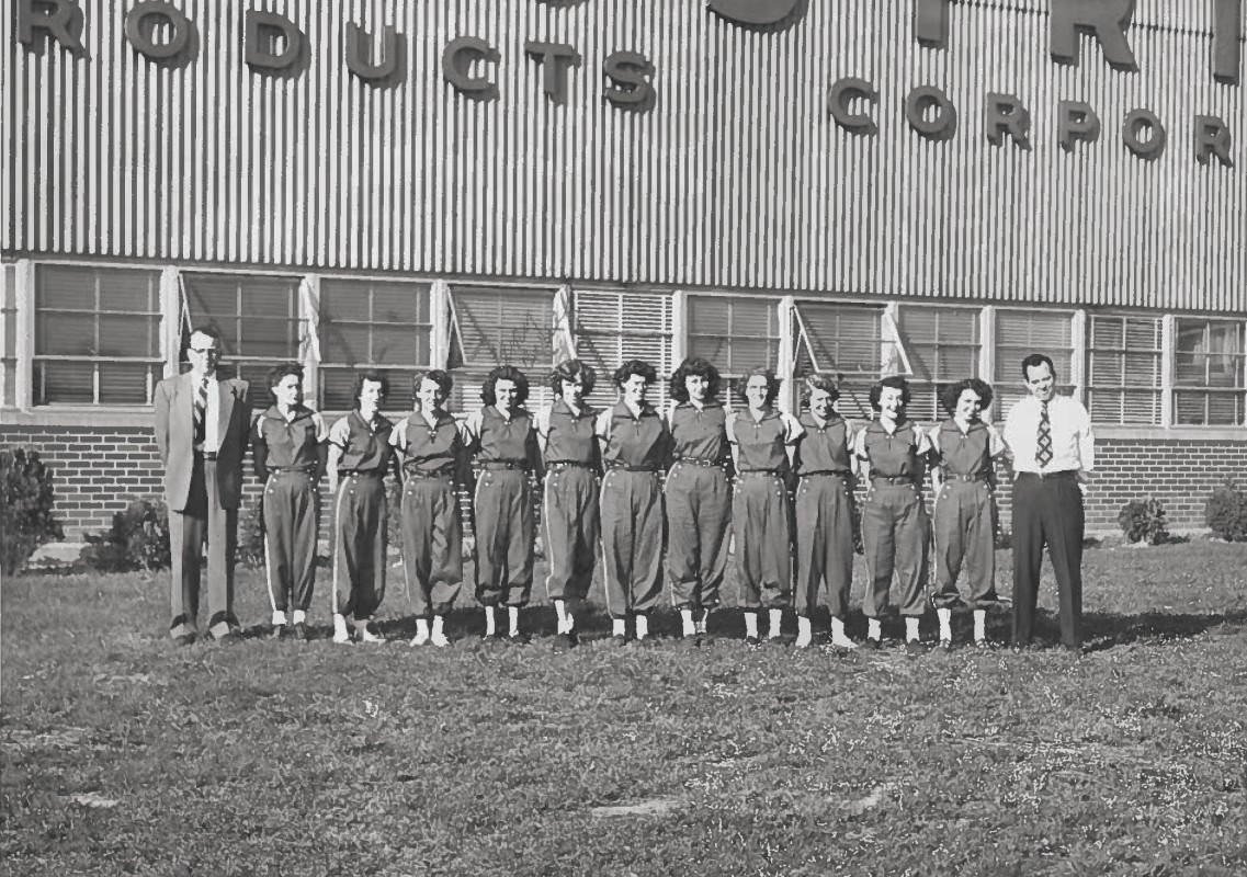Group of factory workers in matching coveralls standing in a line outside a brick building with a 'PRODUCTS CORP' sign above. Two men in suits flank the line at each end.