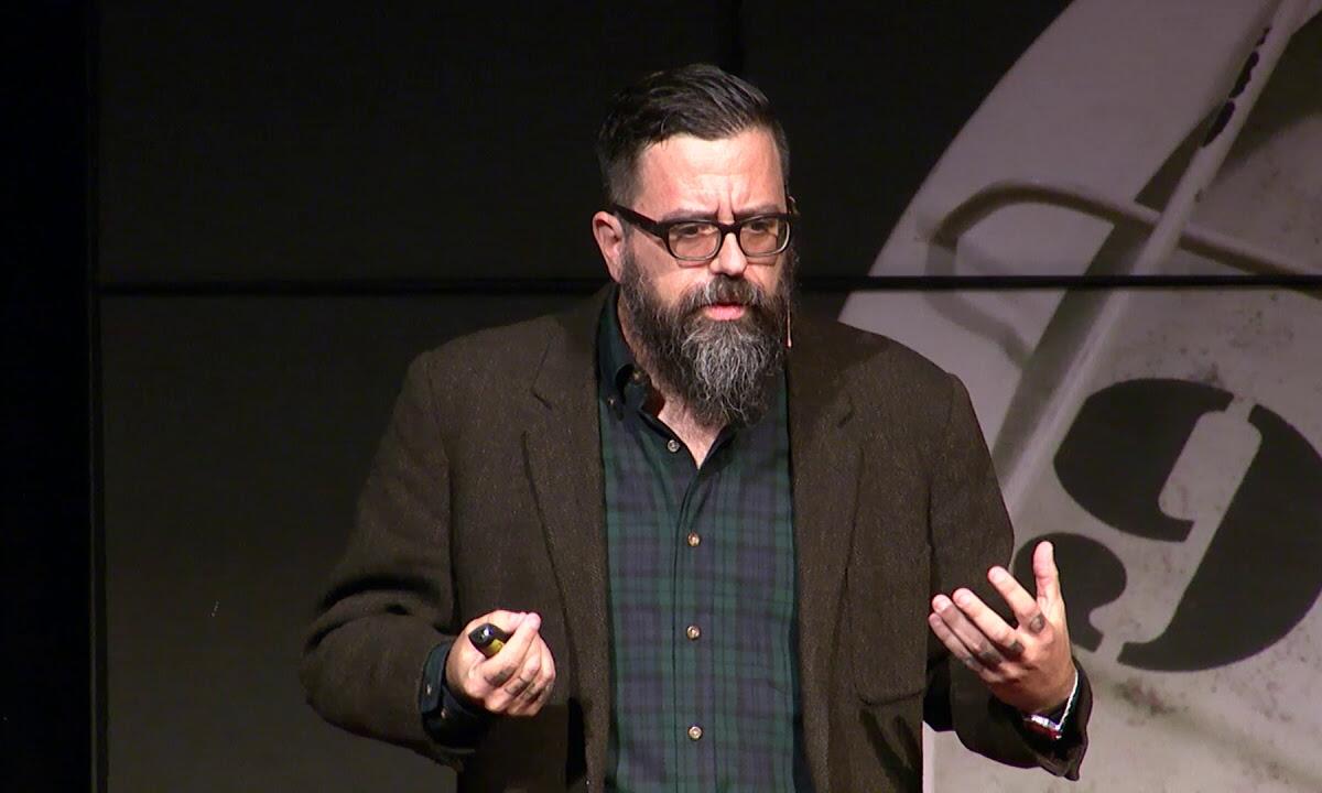 Bearded man with glasses giving a talk on stage, wearing a brown blazer and green plaid shirt, gesturing with his hands.