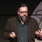 Bearded man with glasses giving a talk on stage, wearing a brown blazer and green plaid shirt, gesturing with his hands.