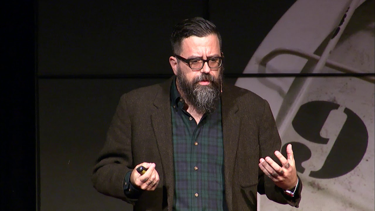 Bearded man with glasses giving a talk on stage, wearing a brown blazer and green plaid shirt, gesturing with his hands.