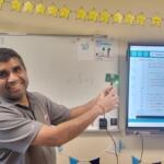 Smiling teacher demonstrates a microcontroller LED project in a classroom, pointing to a small green board beside a whiteboard and a screen showing Arduino code.