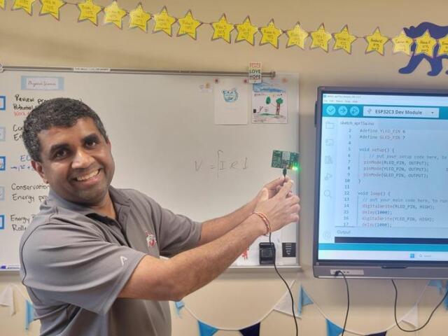 Smiling teacher demonstrates a microcontroller LED project in a classroom, pointing to a small green board beside a whiteboard and a screen showing Arduino code.