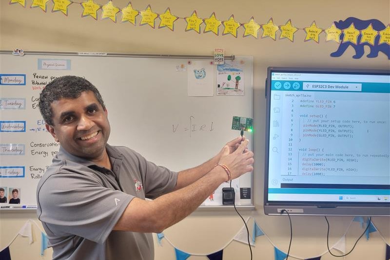 Smiling teacher demonstrates a microcontroller LED project in a classroom, pointing to a small green board beside a whiteboard and a screen showing Arduino code.